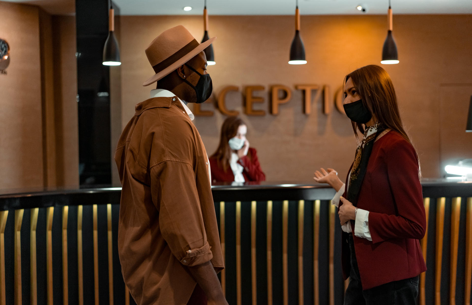 A guest and a hotel staff member wearing masks, talk at the reception desk.
